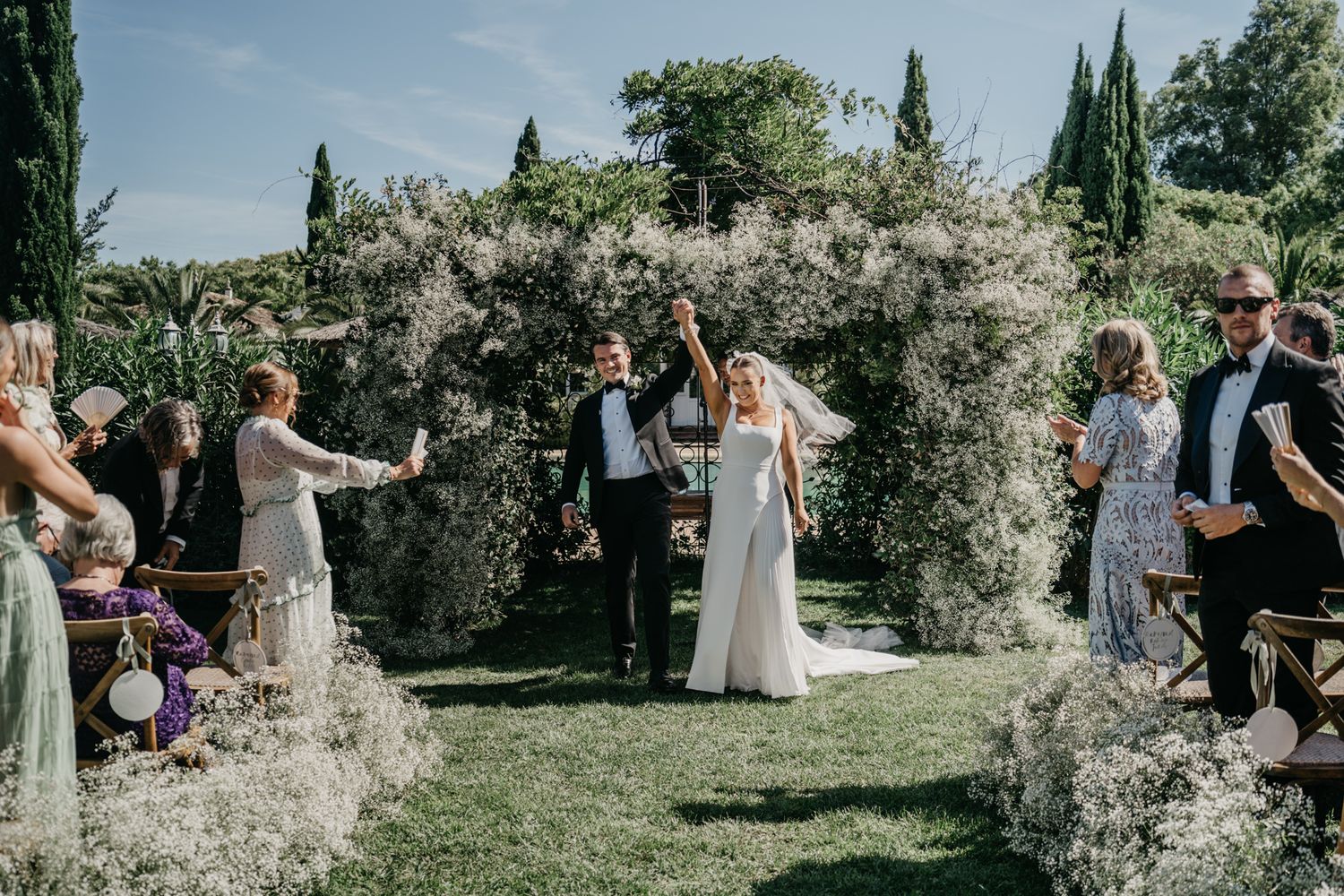 Outdoor wedding ceremony under a floral arch with guests looking on.
