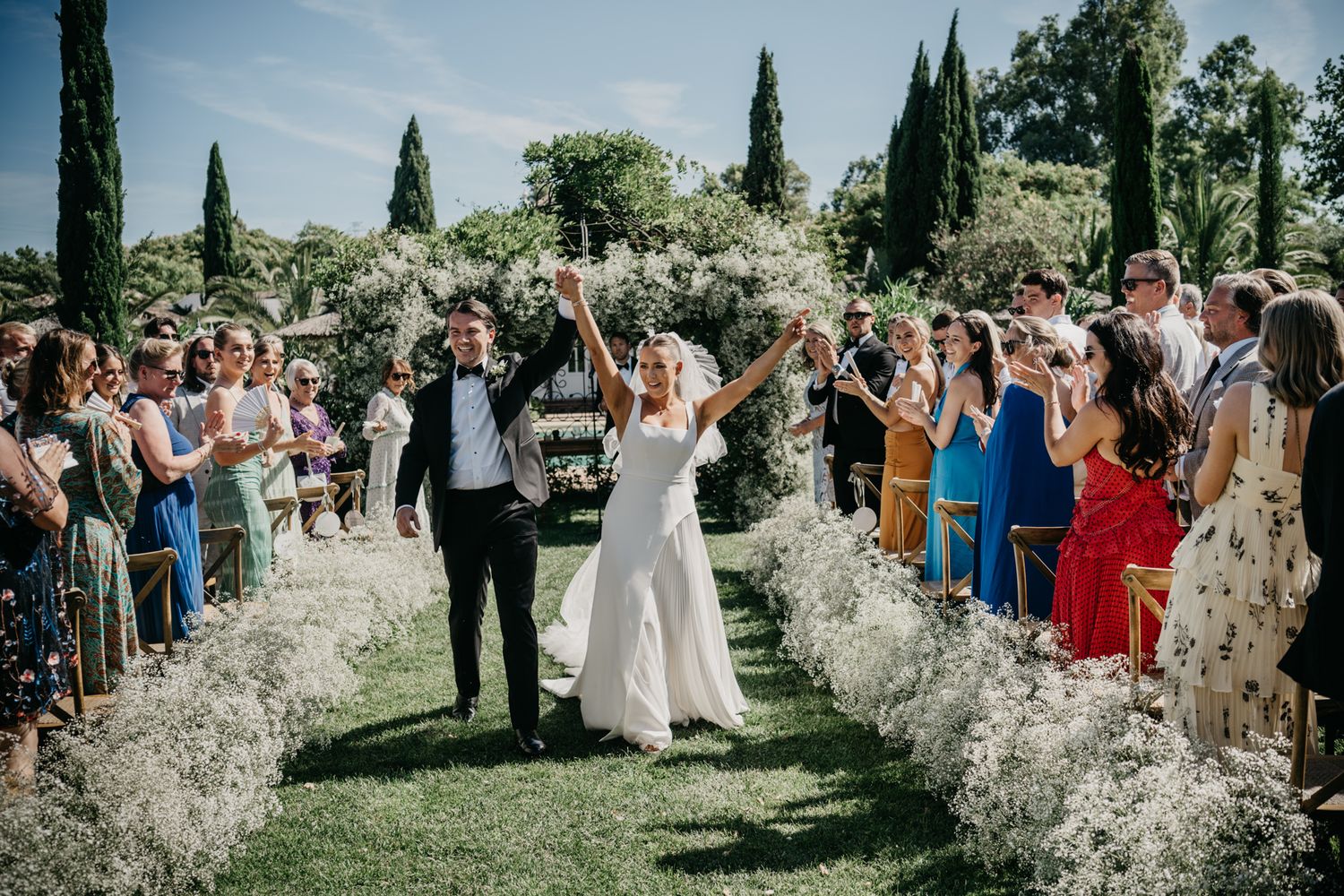 Newlyweds walking down aisle lined with white flowers as guests celebrate.