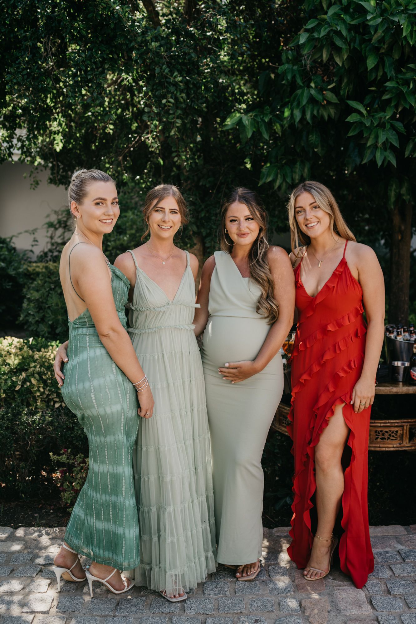 Women in elegant formal dresses posing together at outdoor garden event.