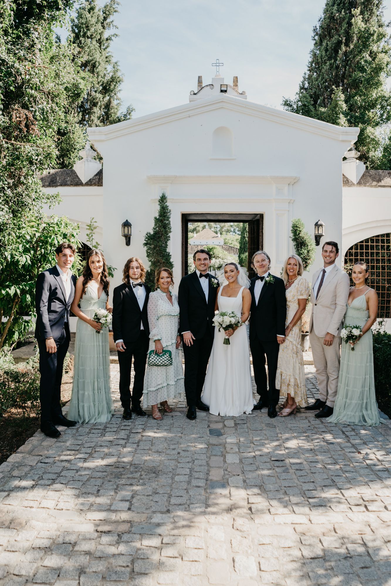 Wedding party in formal attire gathered at white chapel entrance.