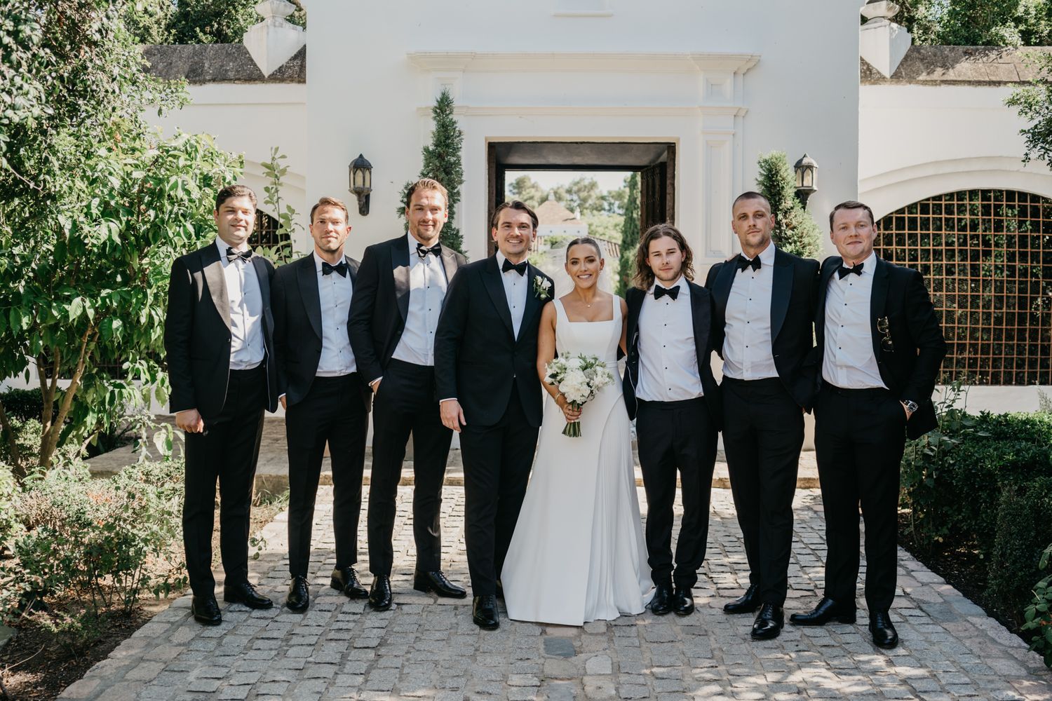 Wedding party in black tie attire posed in front of white chapel entrance.