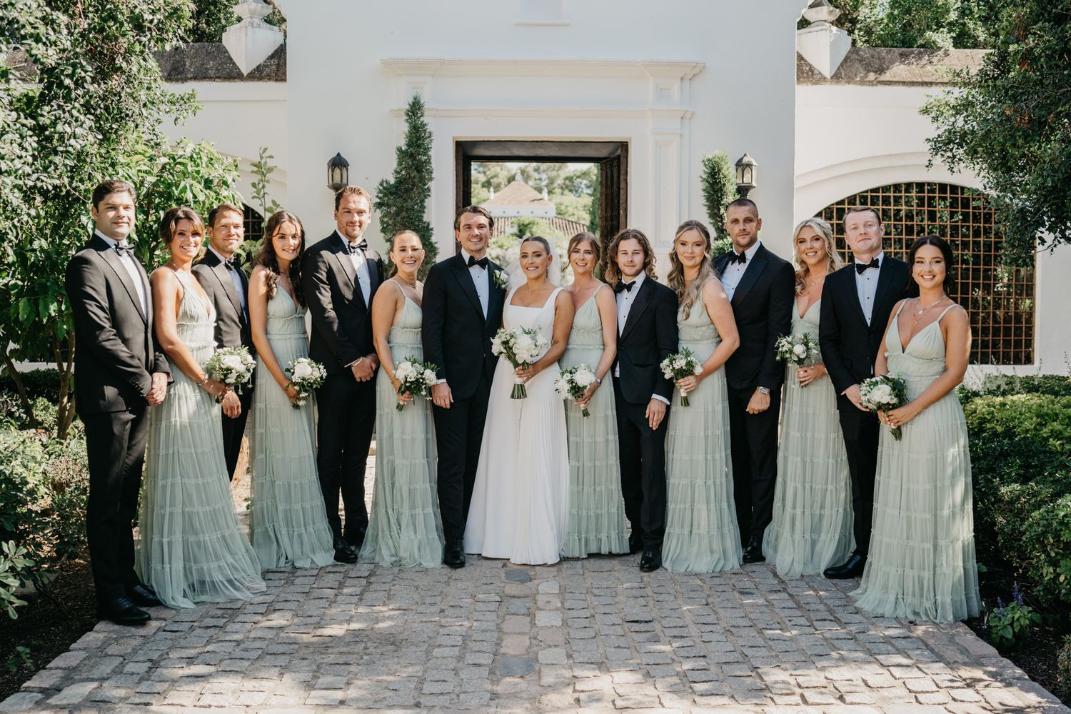 Wedding party gathered outside white Mediterranean building in sage green and formal attire.
