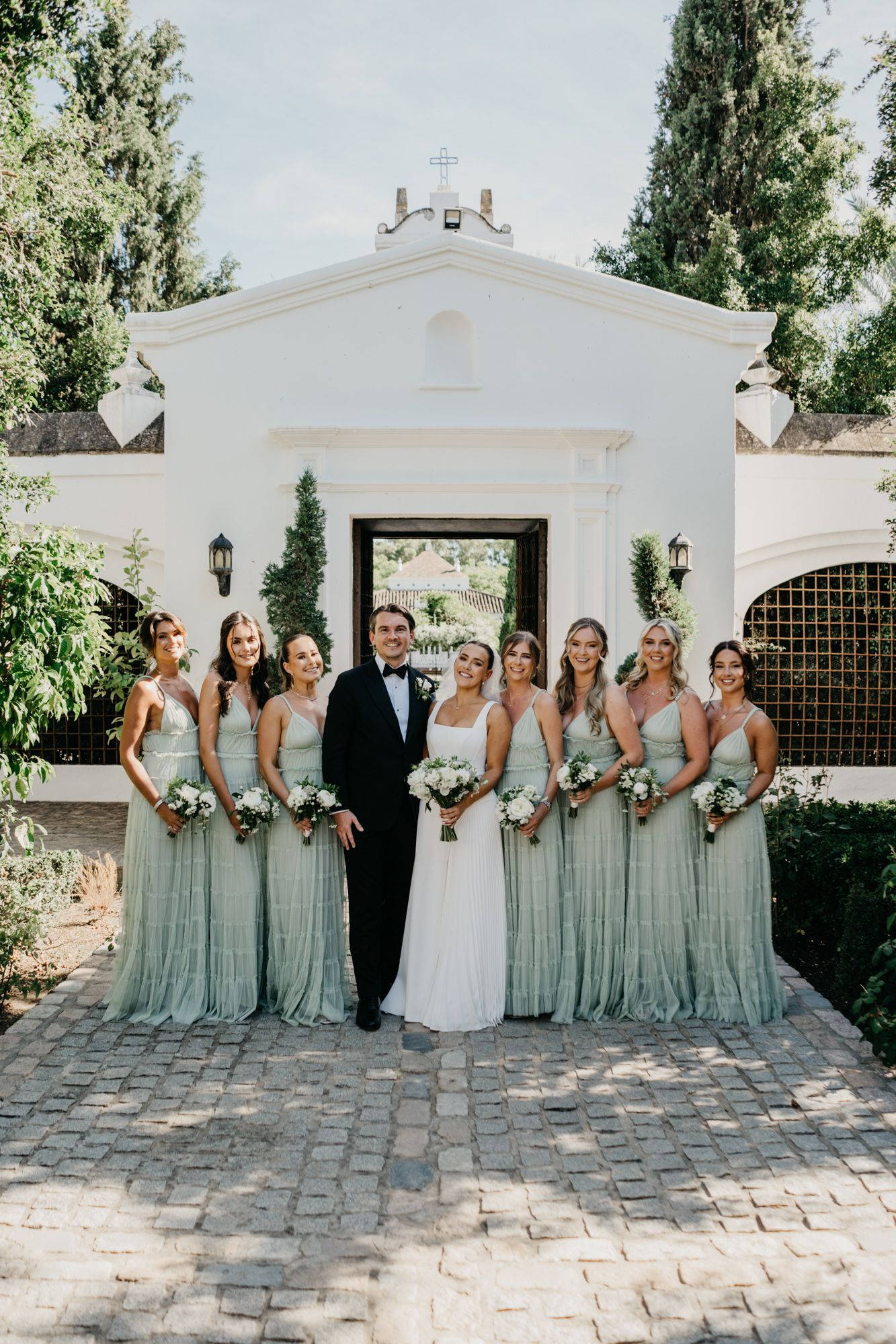 Wedding party poses in front of white Spanish-style building wearing sage green bridesmaid dresses.
