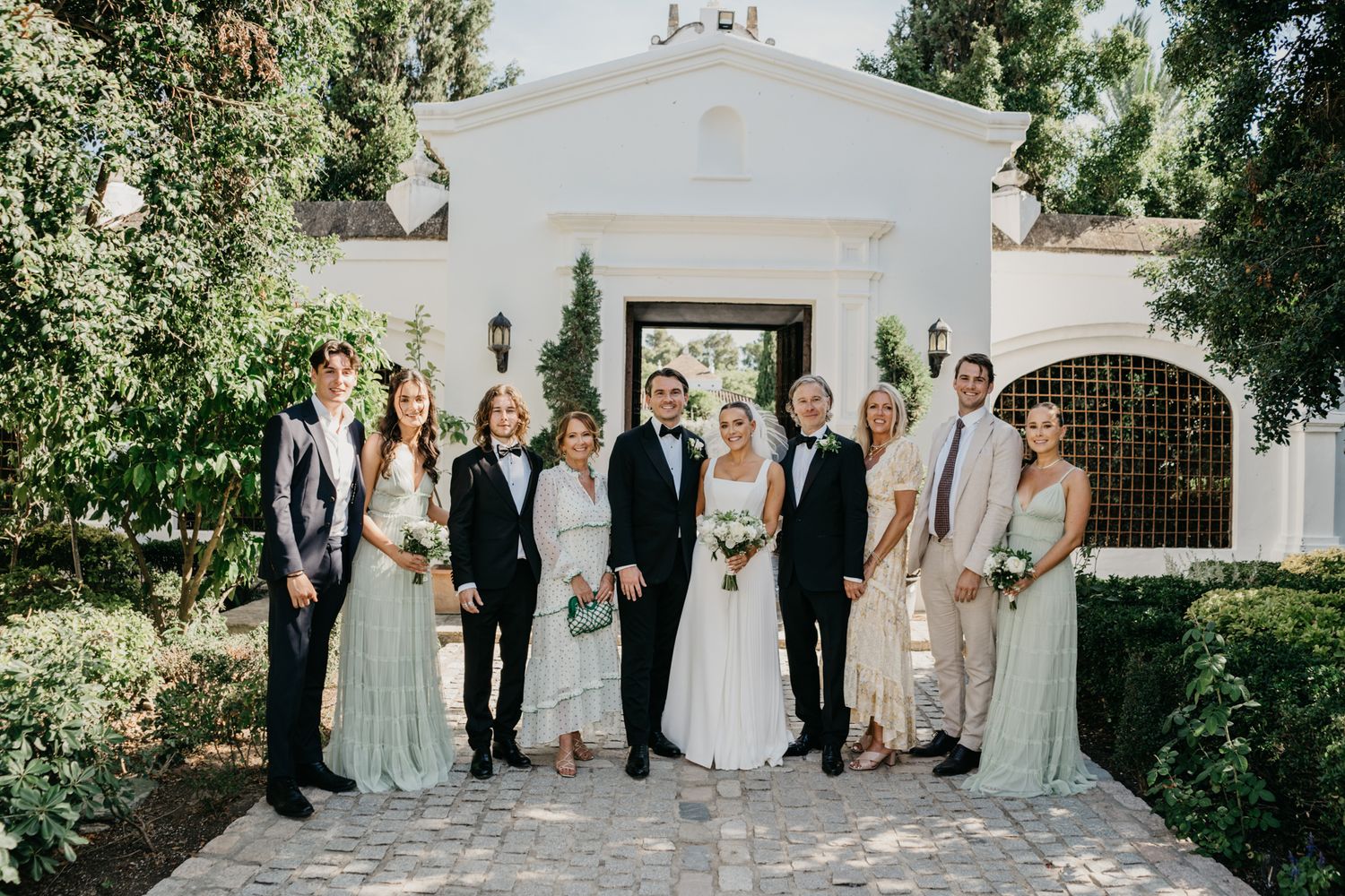 Full wedding party group photo outside white Mediterranean-style venue with greenery.