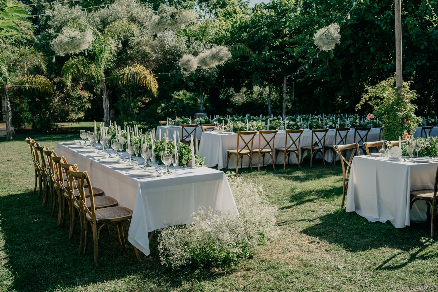 Outdoor wedding reception setup with white tablecloths and wooden cross-back chairs.