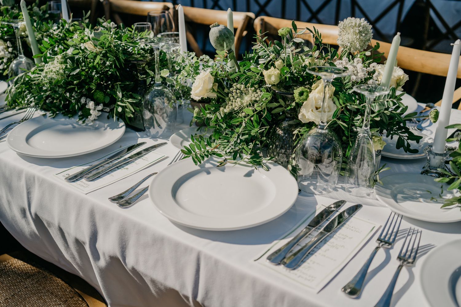 Elegant wedding table setting with white linens, plates, and fresh greenery centerpieces.