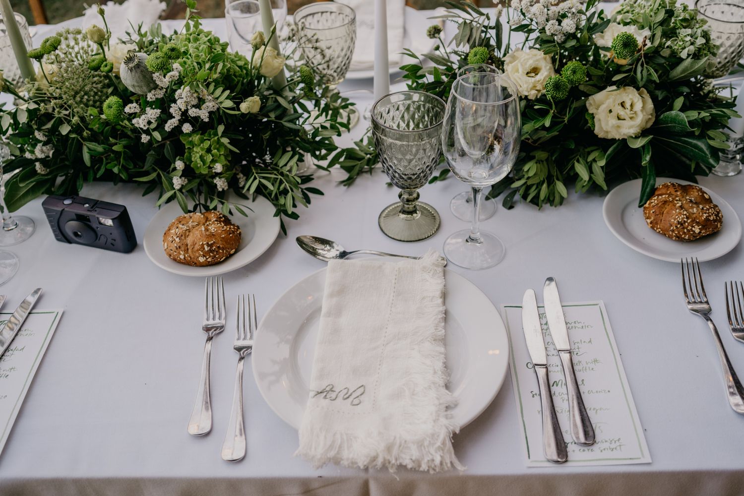 Detailed view of wedding place setting with white napkin, crystal stemware, and fresh greenery.