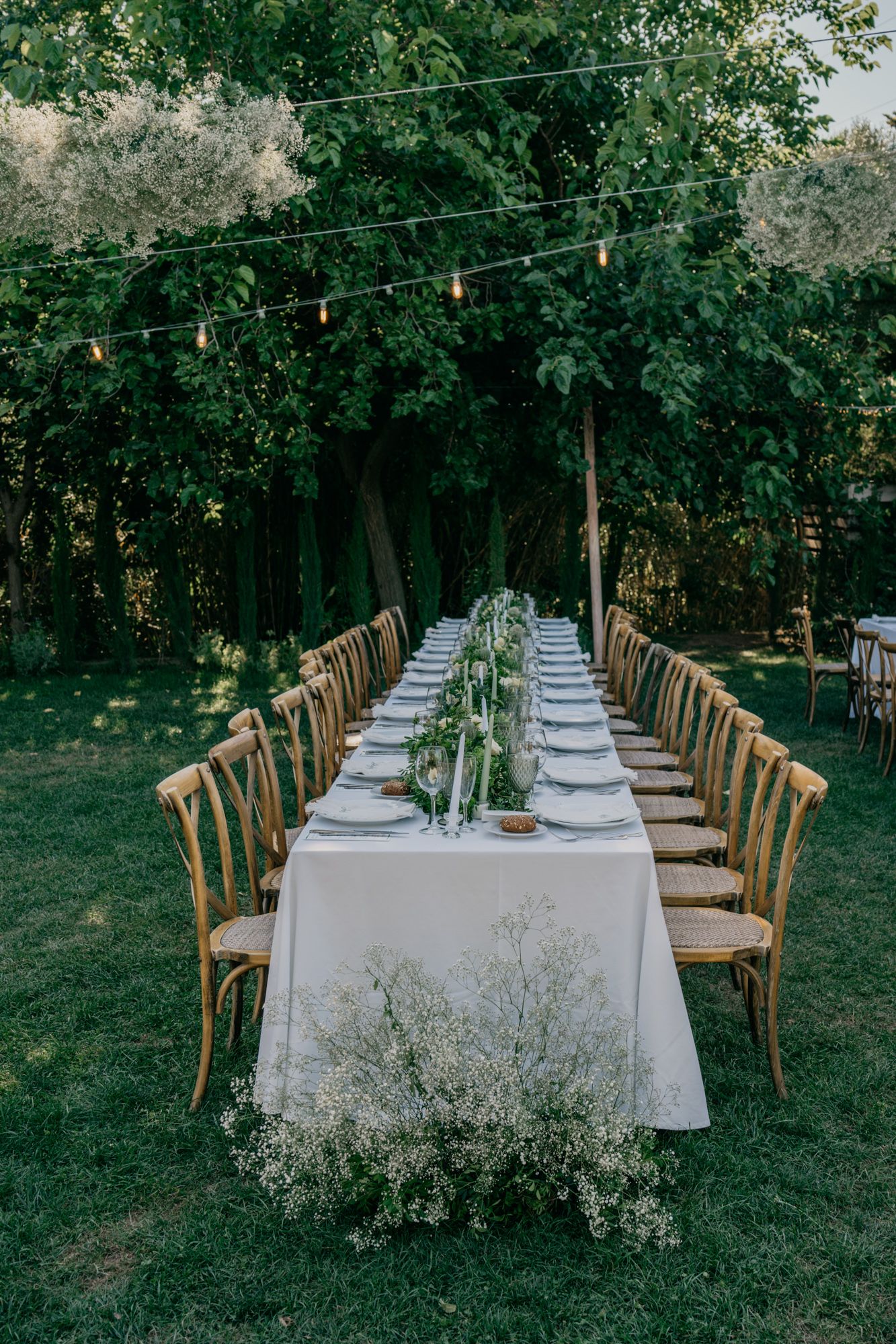 Outdoor wedding reception setup with white tablecloth and wooden chairs under string lights.