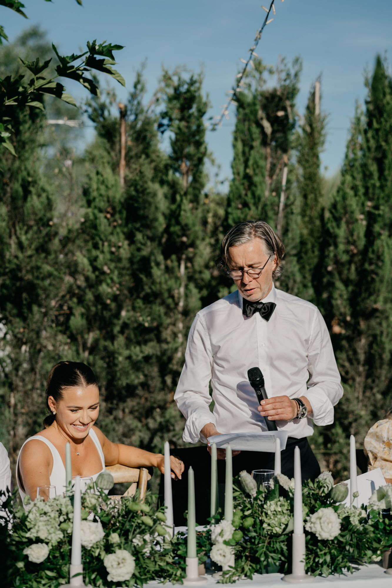 People at a formal outdoor dining table decorated with white floral centerpieces and candles.