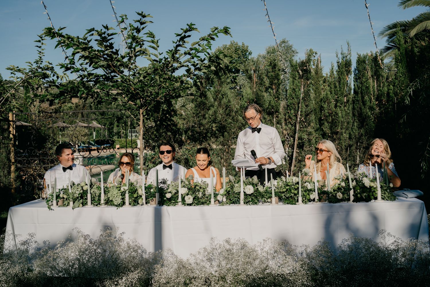 Wedding party seated at a long white table decorated with greenery outdoors.