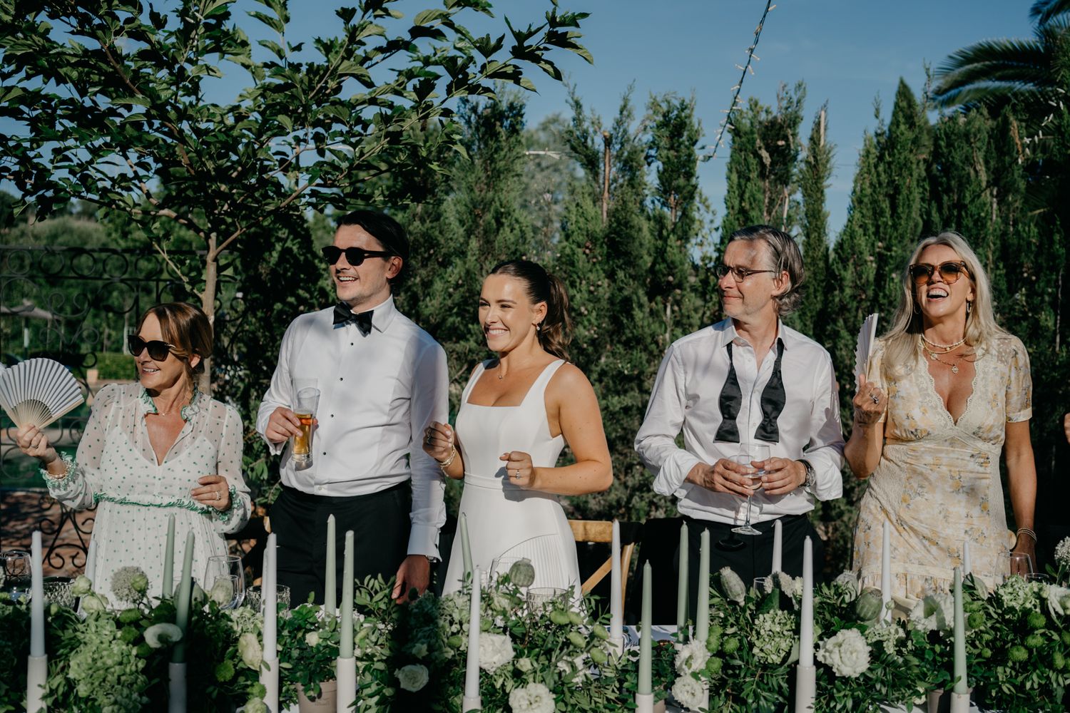 Wedding party members standing behind a decorated table in an outdoor garden setting.