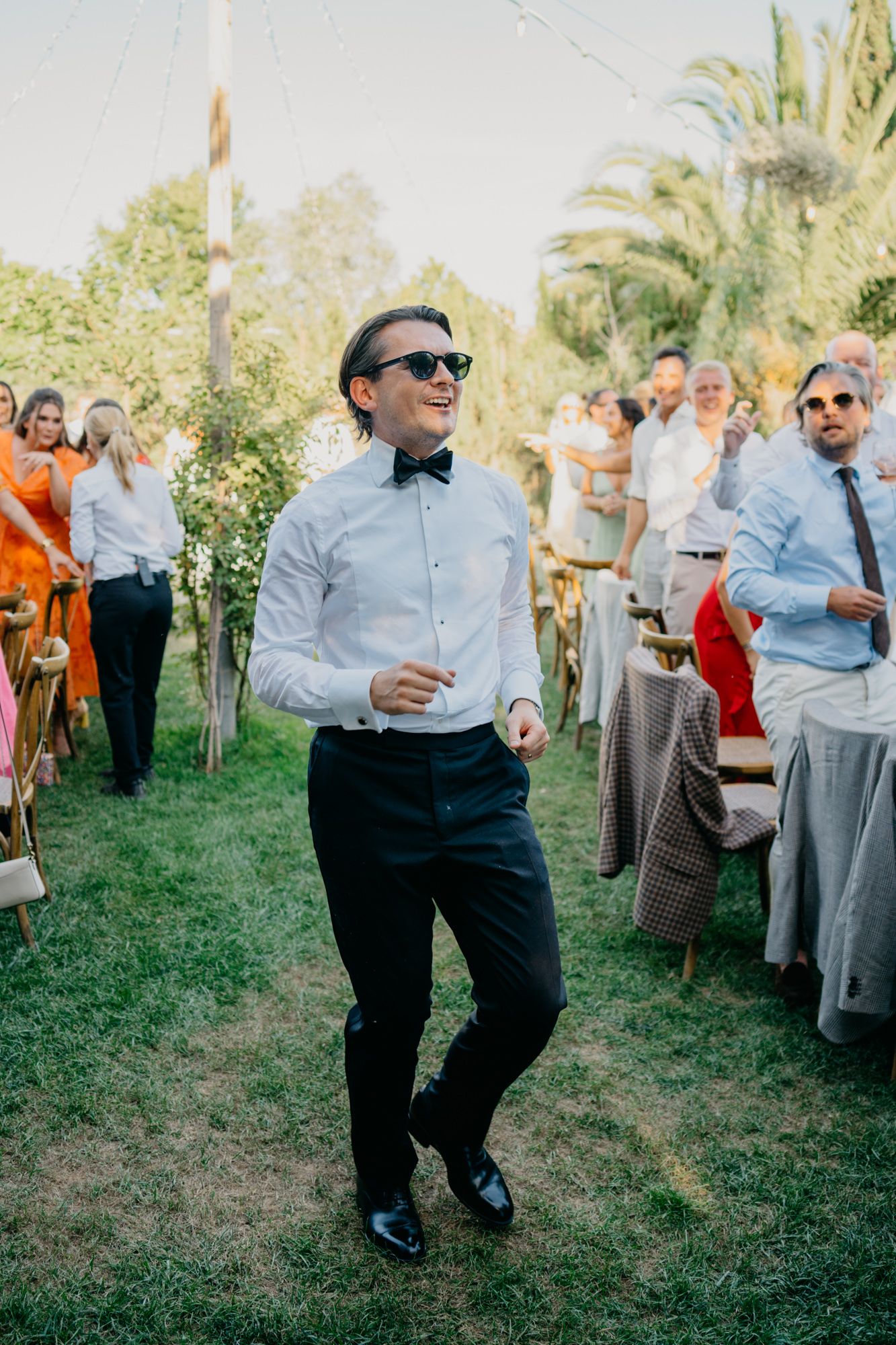 Stylishly dressed wedding guest in sunglasses dances on grass during outdoor celebration.