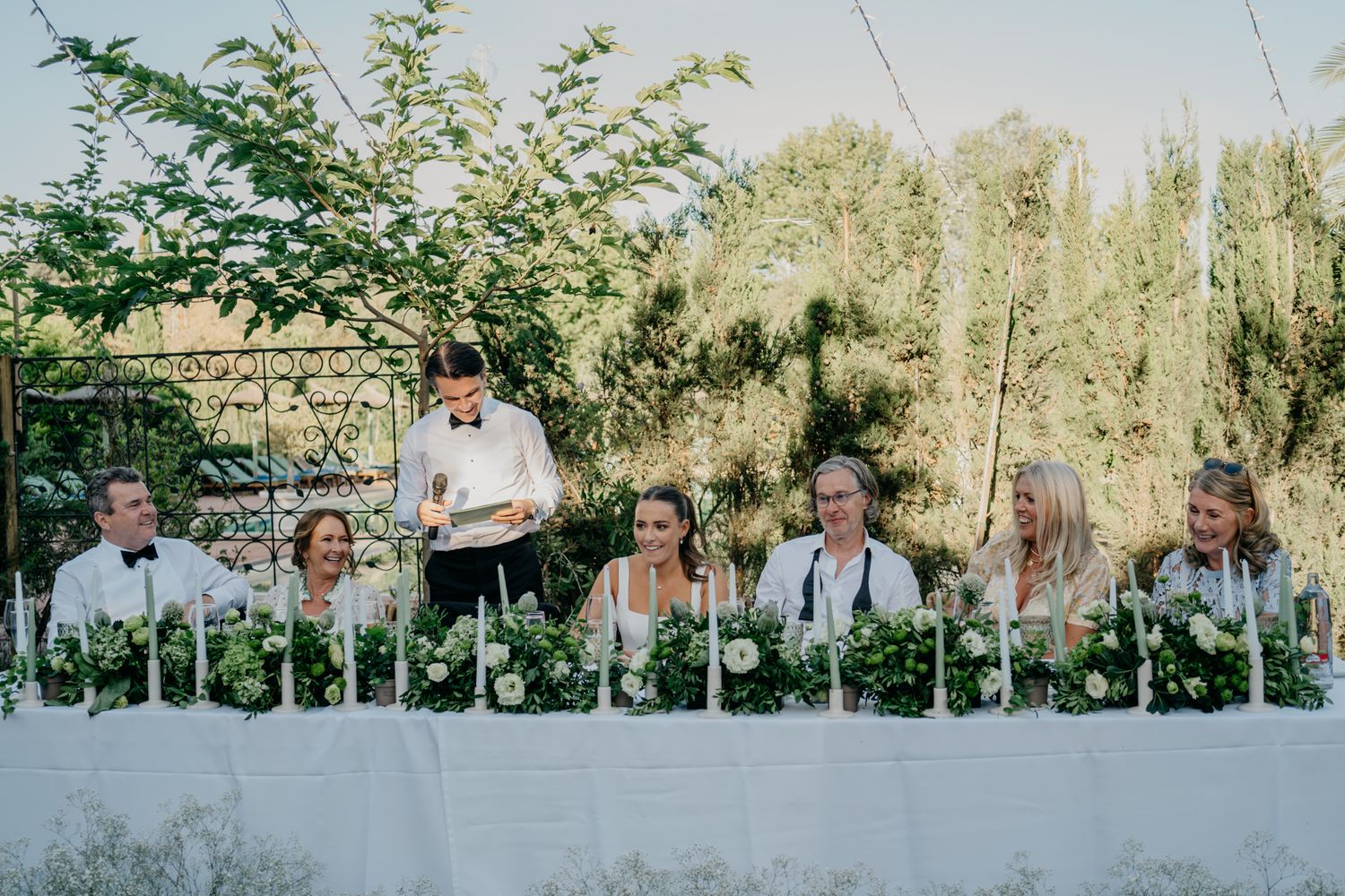 Wedding party members seated at long white table under garden pergola.