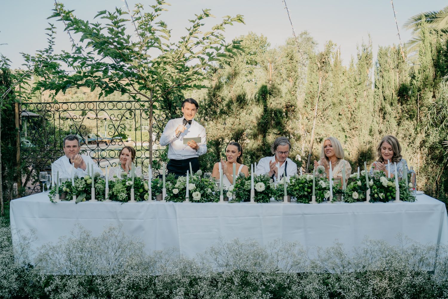 Wedding reception table setup outdoors with white linens and greenery decorations.