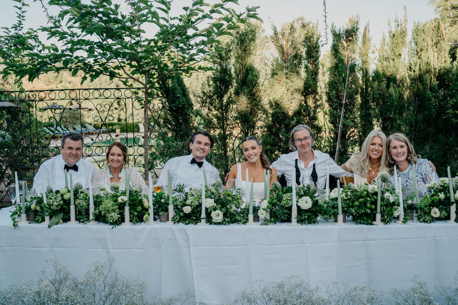 Wedding party seated at long white table decorated with greenery.