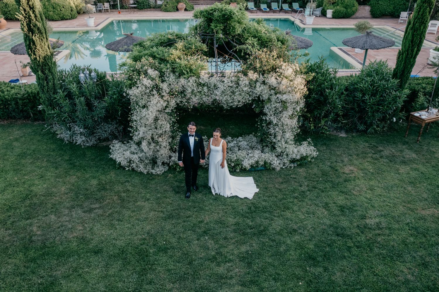 Aerial view of a wedding ceremony setup with white floral arch and pool in the background.