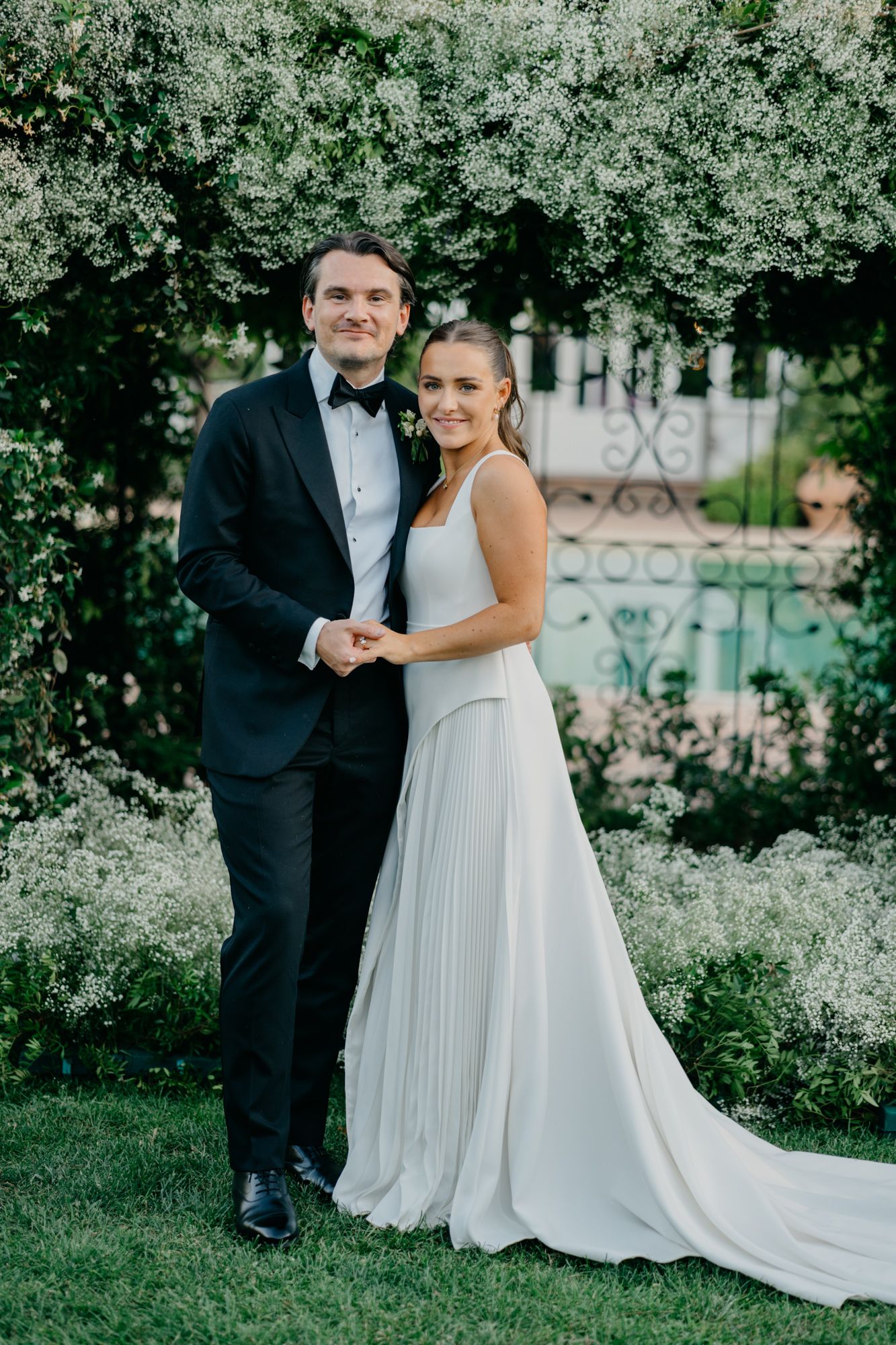Elegant couple posing in garden setting with bride wearing flowing white gown.