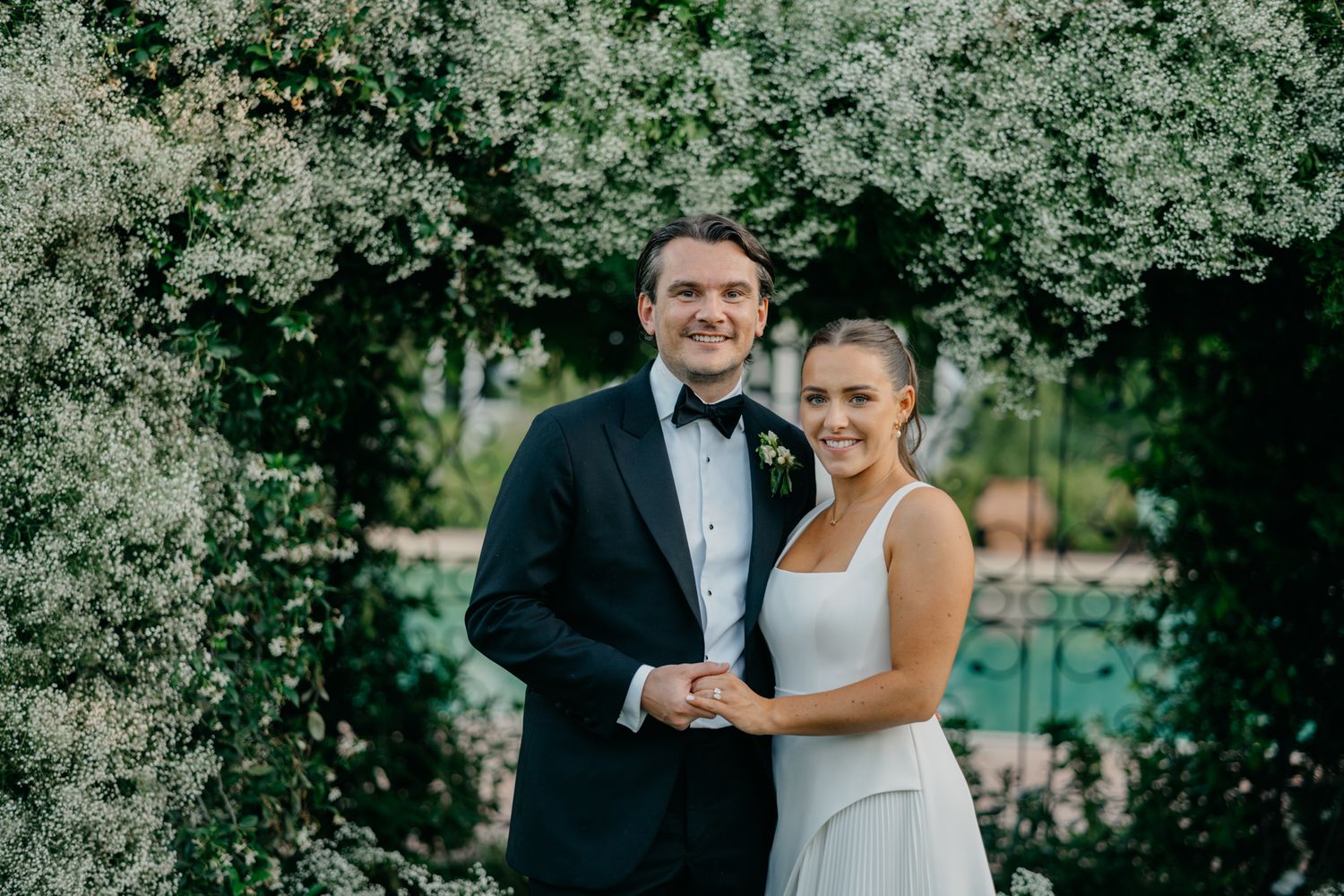 Newlywed couple smiling in garden setting with lush greenery backdrop.
