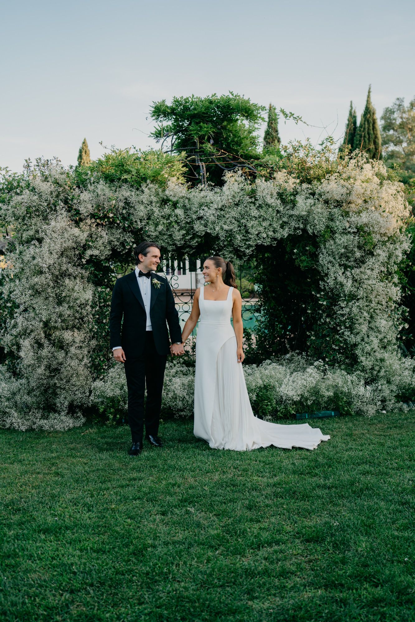 Couple standing under floral arch during outdoor garden wedding ceremony.