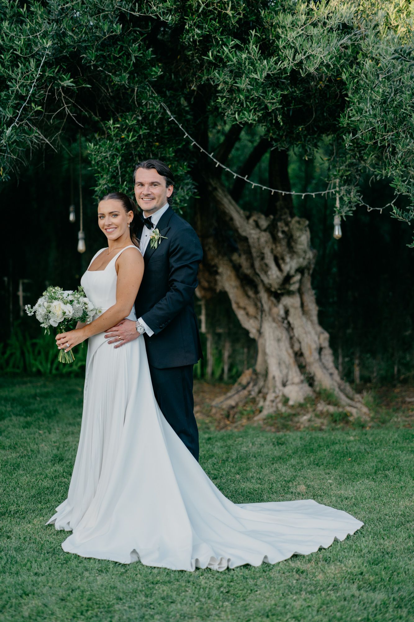 Wedding portrait with couple posing by ancient olive tree with string lights.