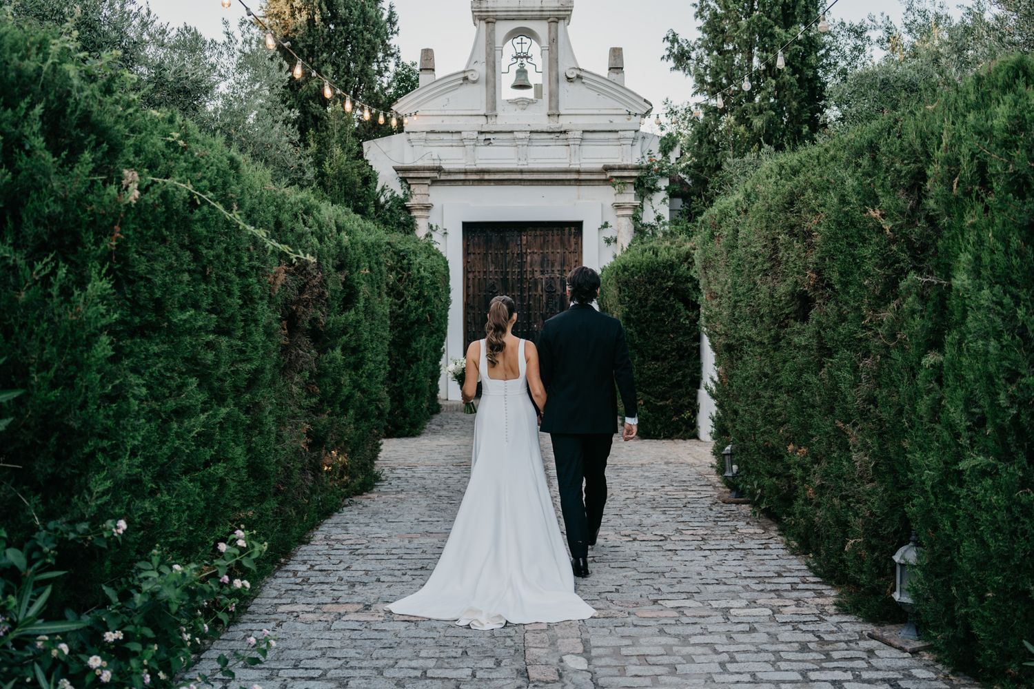 Couple walking towards ornate white chapel entrance through manicured hedge path.