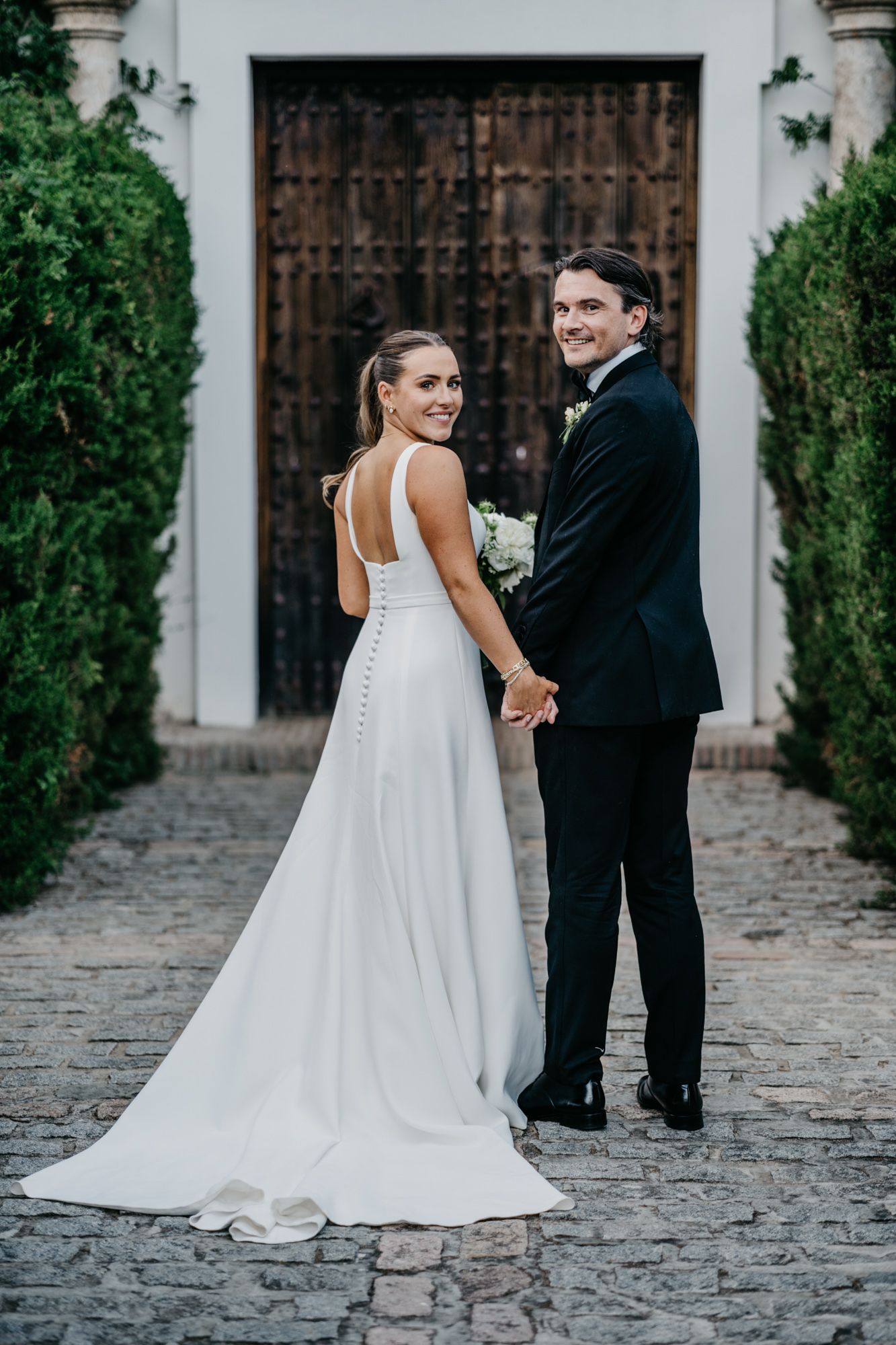 Couple posing by ornate wooden door with bride's dress trailing on cobblestone path.