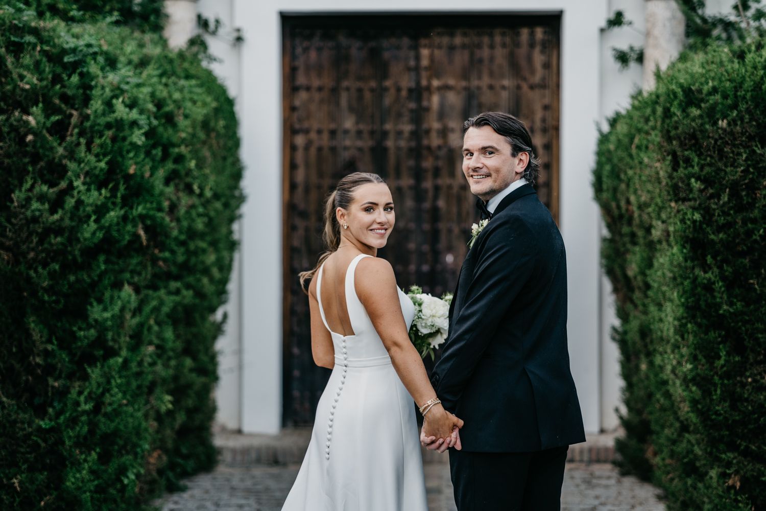 Wedding couple sharing joyful moment in front of rustic doorway with greenery.