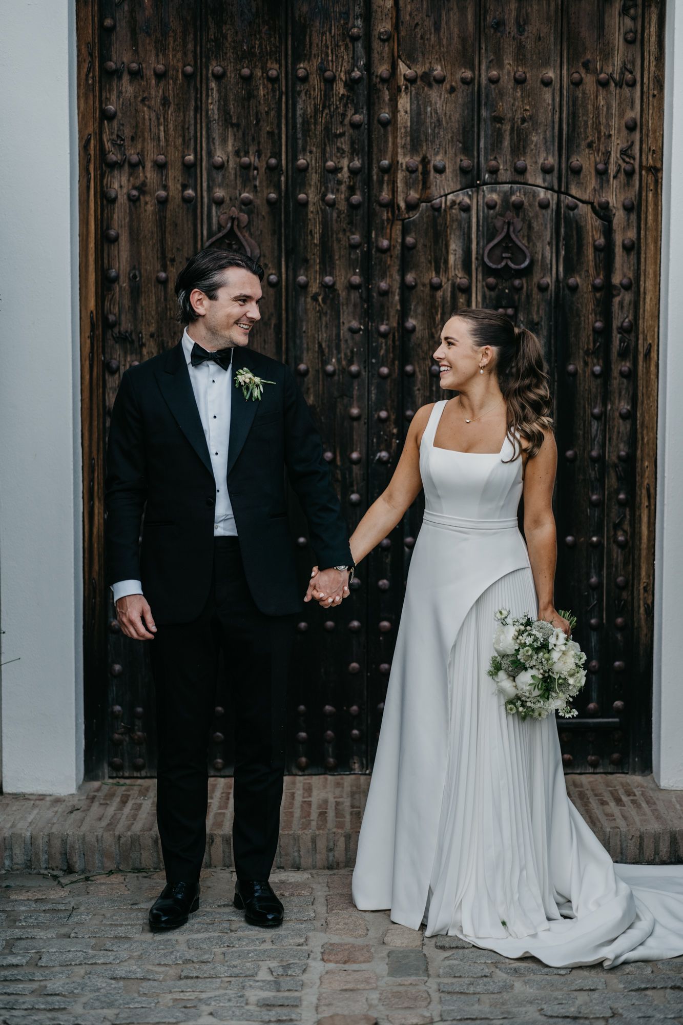 Newlyweds share a joyful moment in front of a rustic wooden door, the bride holding a delicate white bouquet.