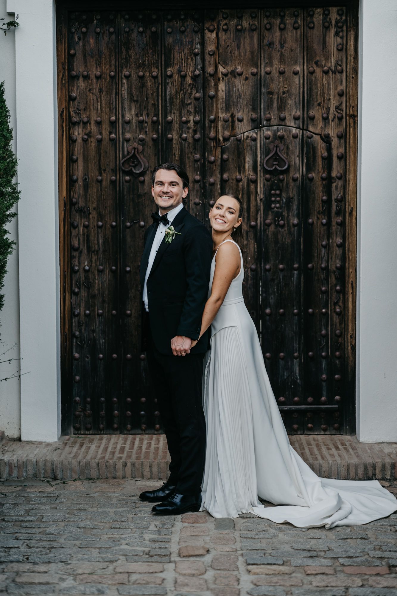 Wedding couple poses against ornate wooden door in formal attire.