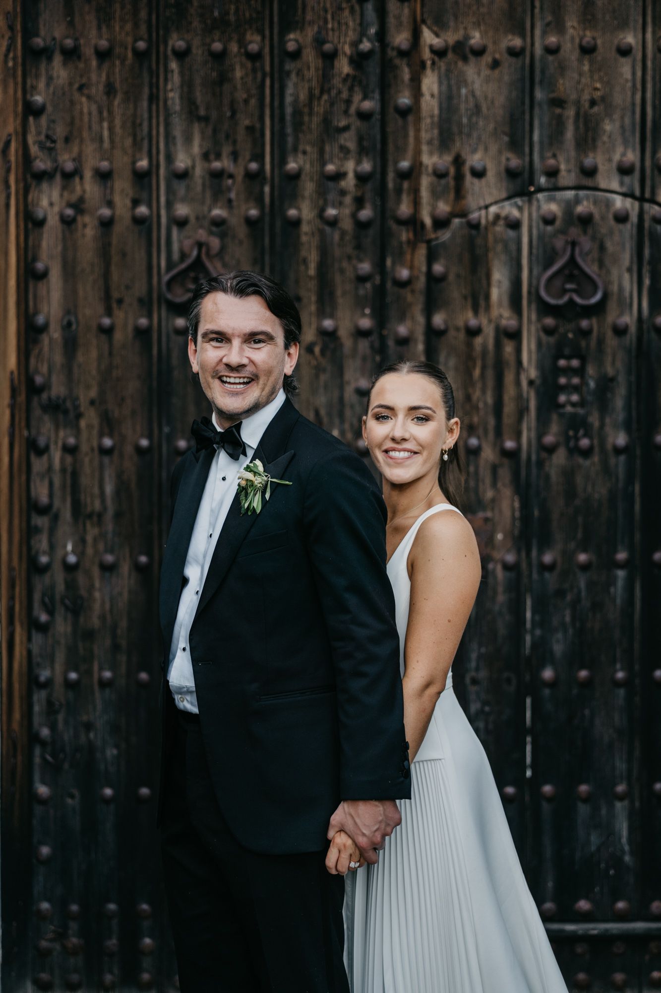 Wedding couple poses against ornate wooden door backdrop in classic black and white attire.