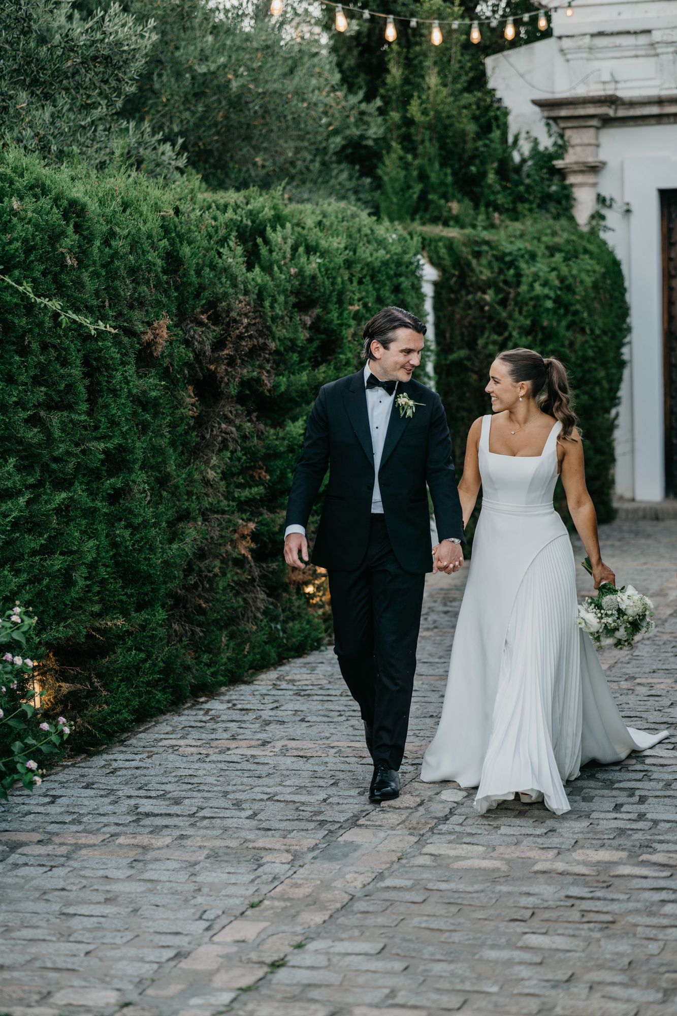 Wedding couple strolls hand-in-hand along cobblestone path surrounded by garden greenery.
