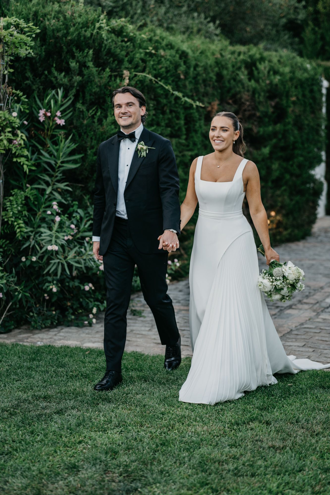 Newlyweds walk together on garden path with bride carrying white floral bouquet.