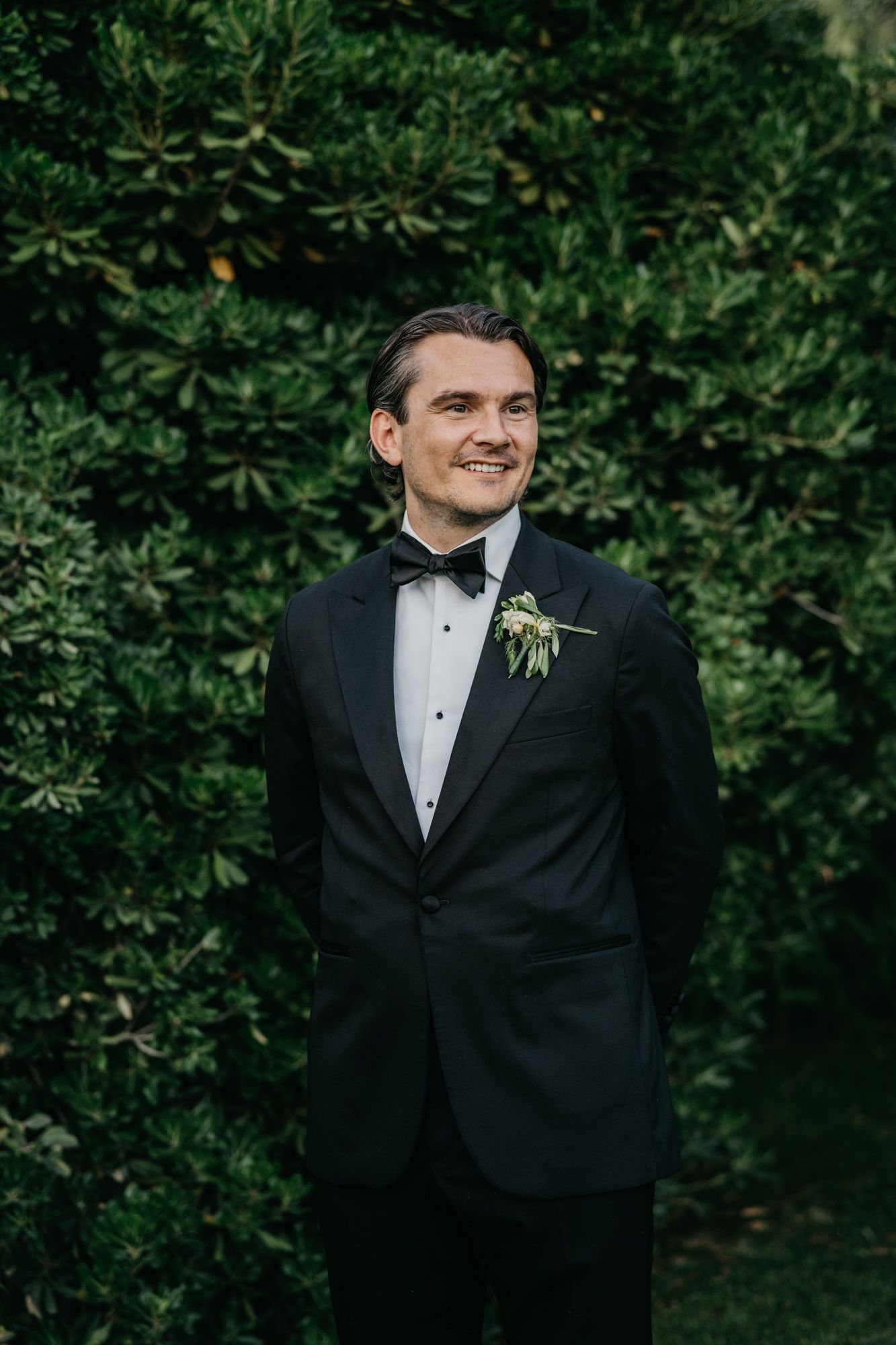 Groom in classic black tuxedo poses against lush garden backdrop.