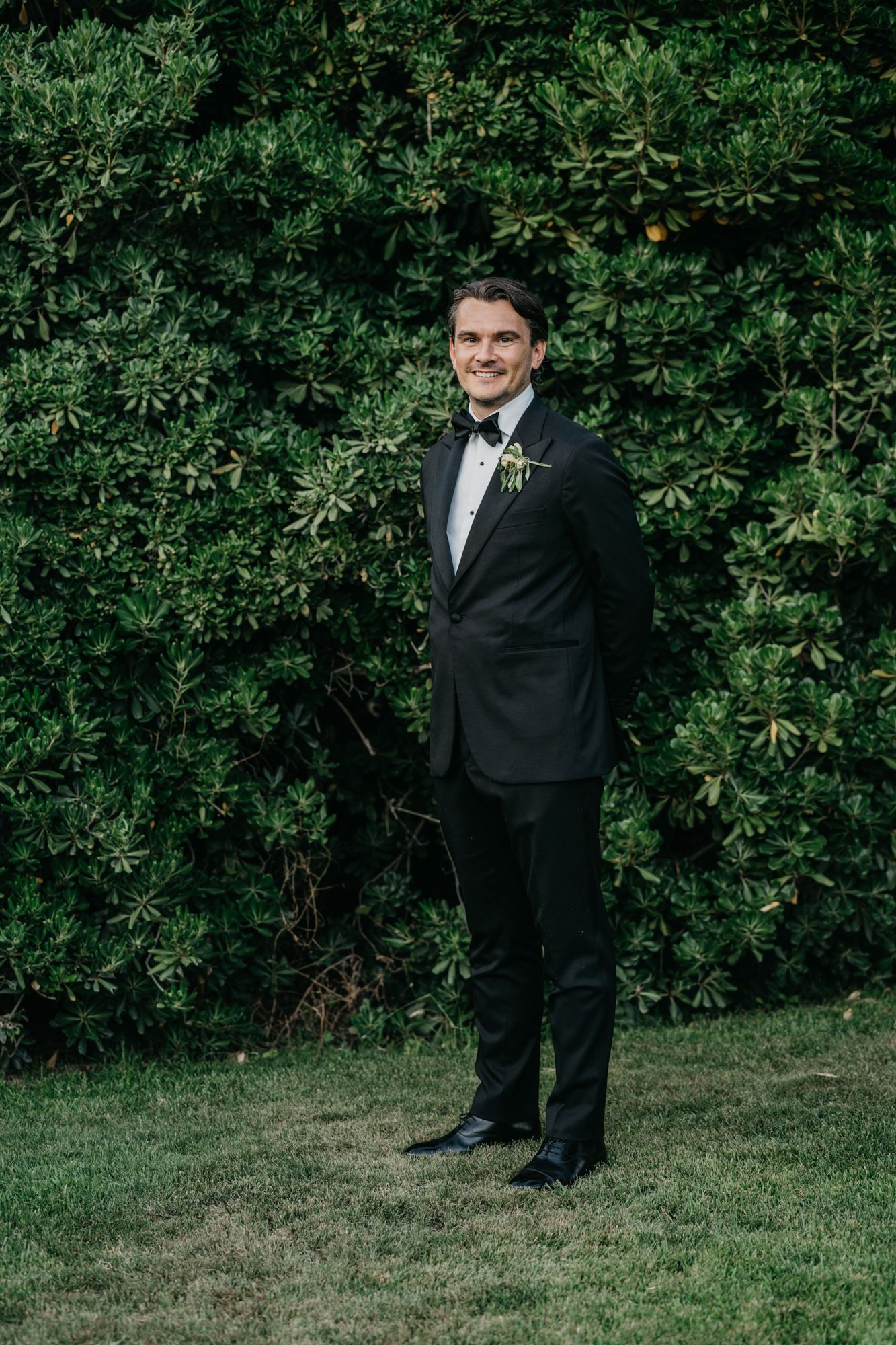 Groom in classic black tuxedo and bowtie poses against lush green garden backdrop.