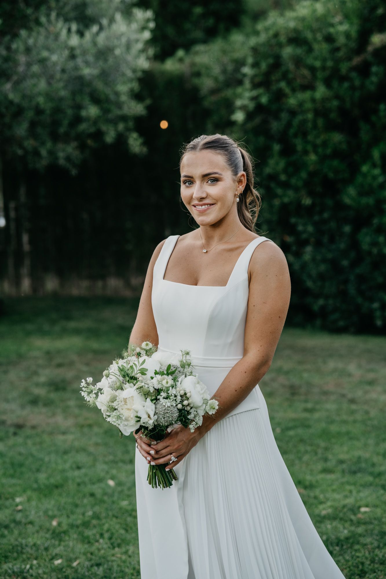 Bride in elegant white dress holds bouquet of white flowers in outdoor garden setting.