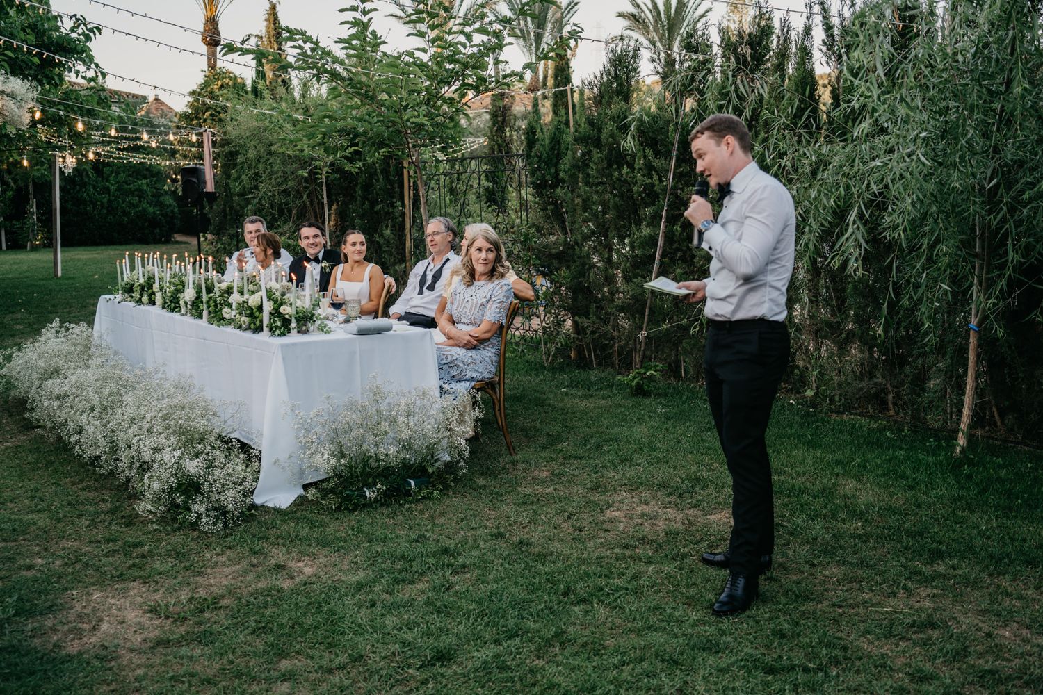Wedding reception setup with long white table and string lights in garden setting.