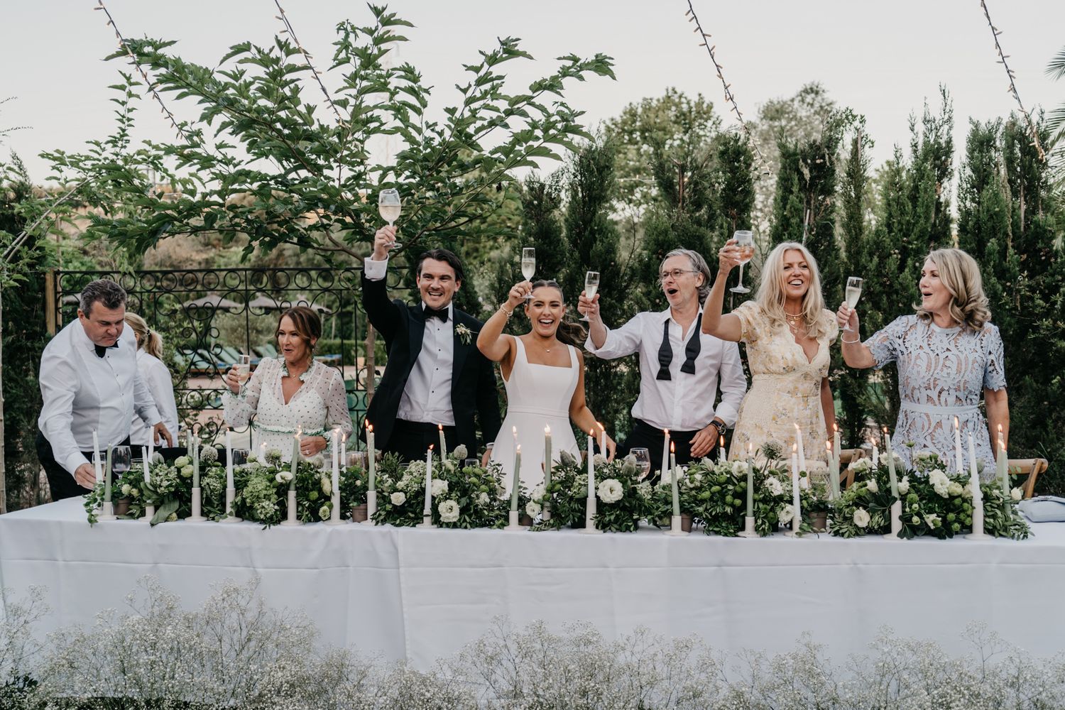 Wedding party poses together on white benches in a garden setting with string lights overhead.