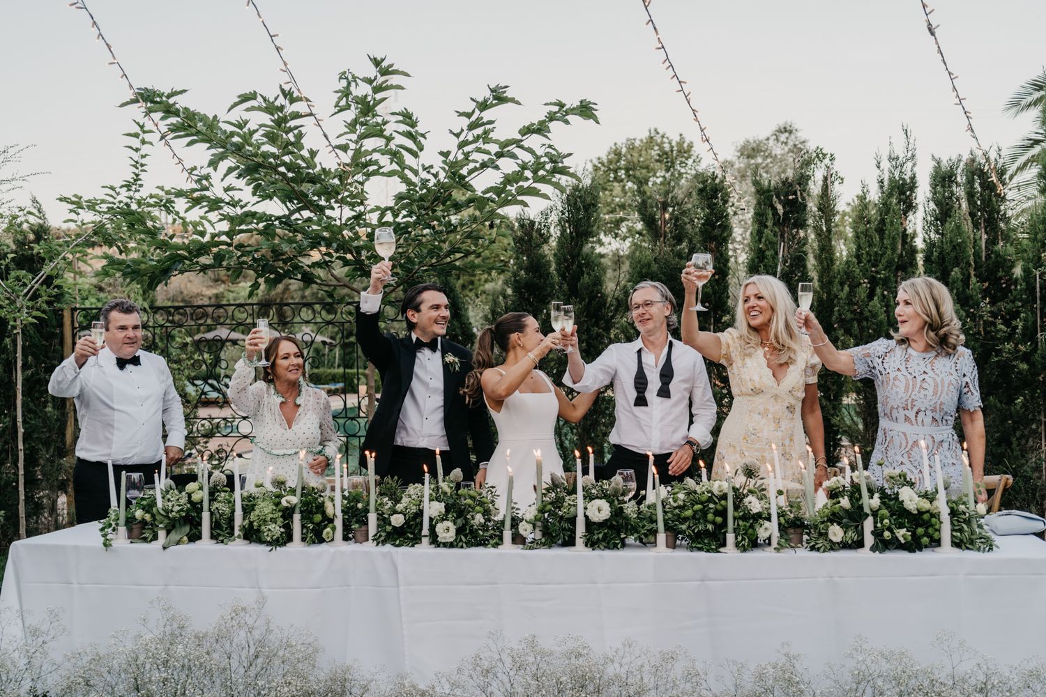 Wedding party stands behind long white table at outdoor garden reception.