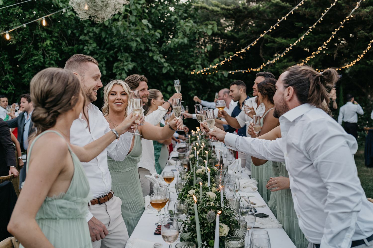 Wedding guests toast across long tables decorated with greenery under twinkling string lights.