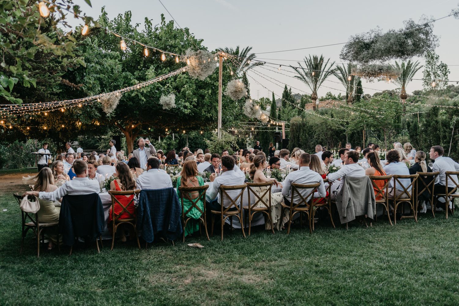 Outdoor wedding reception with guests seated at long tables under string lights.