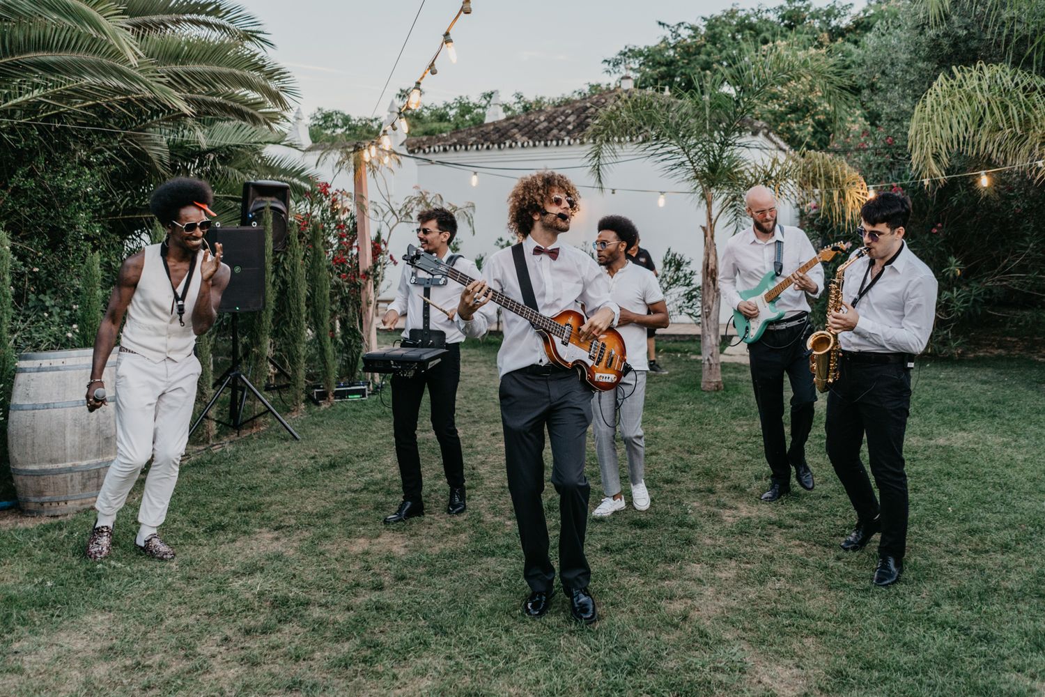 Wedding band performs outdoors with string instruments and casual attire among palm trees.
