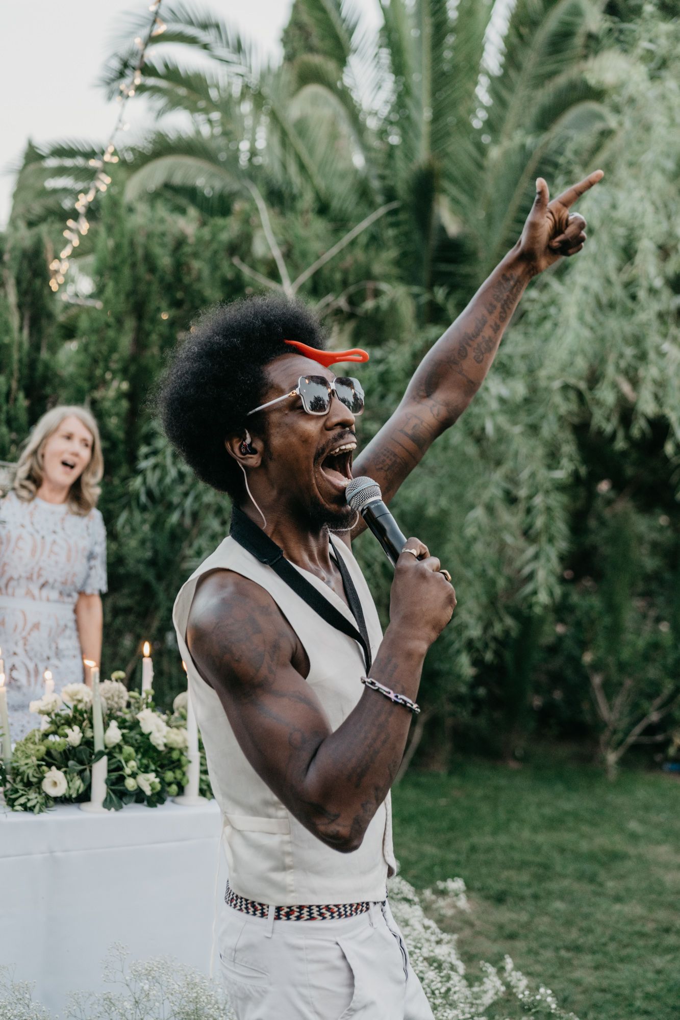 Energetic performer in white outfit sings with passion at outdoor garden celebration.