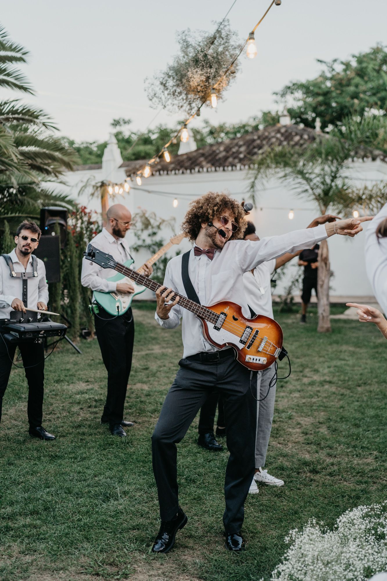 Musicians perform with guitars at outdoor event beneath string lights and palm trees.