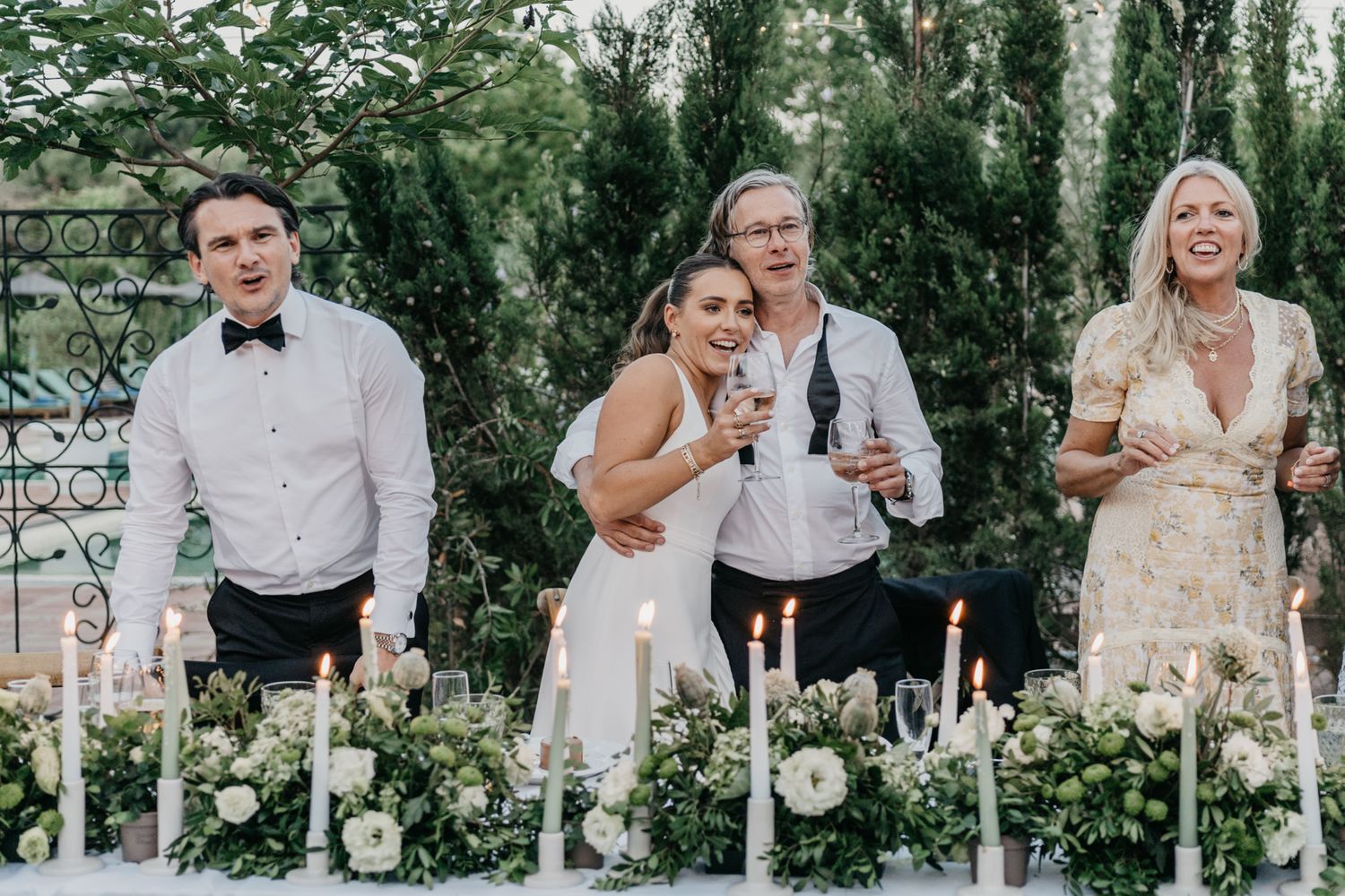 Wedding party laughs together at decorated outdoor reception table.