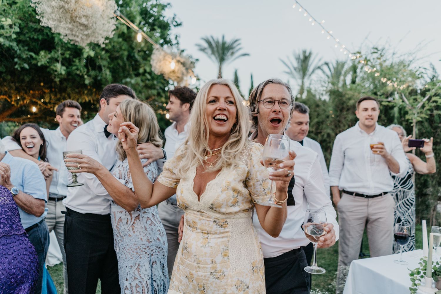 Wedding guests laughing and celebrating with drinks at an outdoor garden reception.