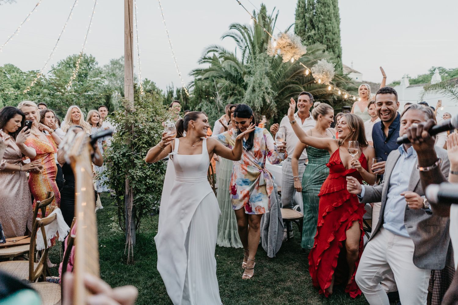 Wedding celebration in full swing as guests dance and cheer at an outdoor garden venue.