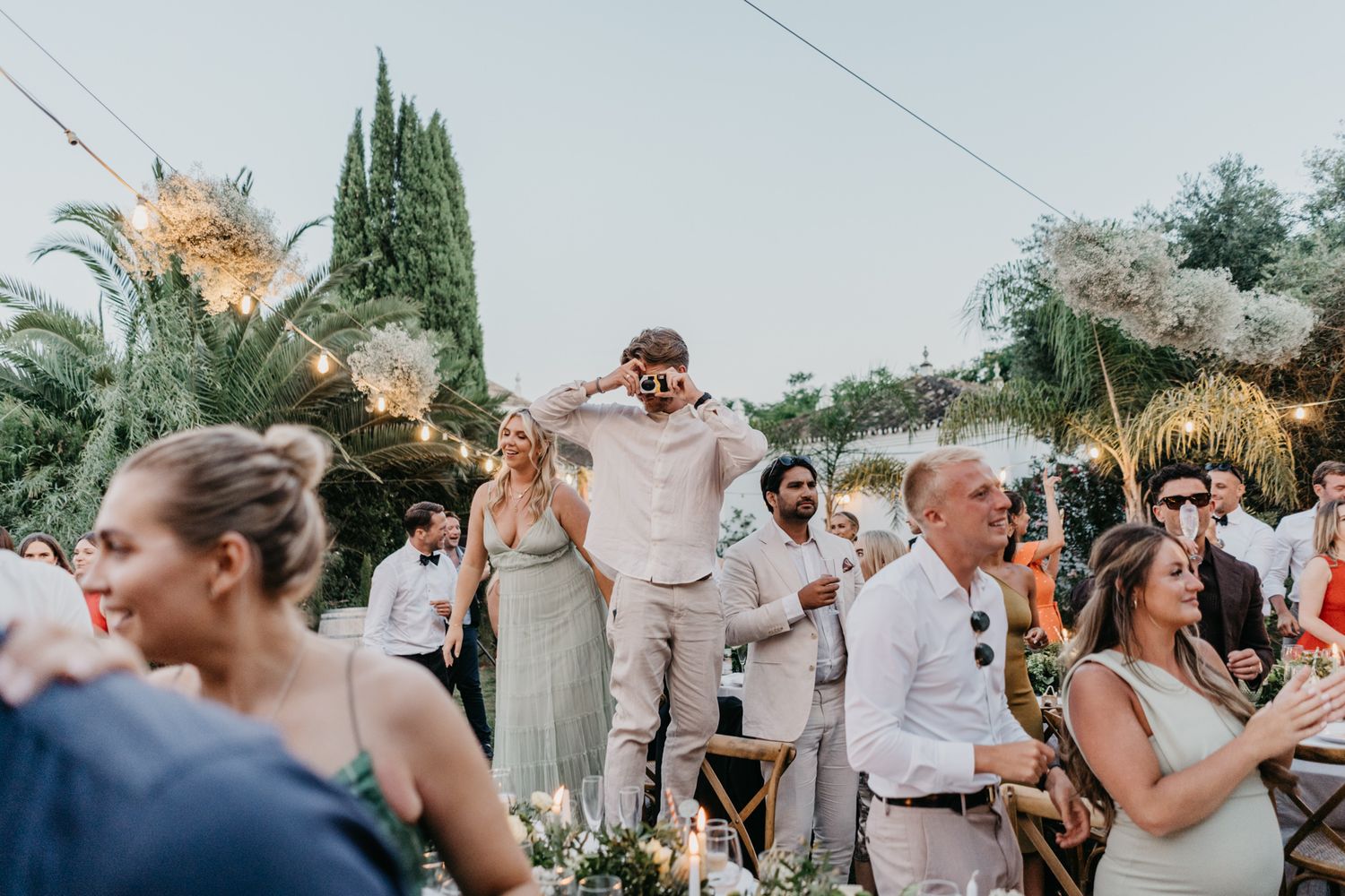 Wedding guests dancing and celebrating at outdoor reception with string lights and palm trees.