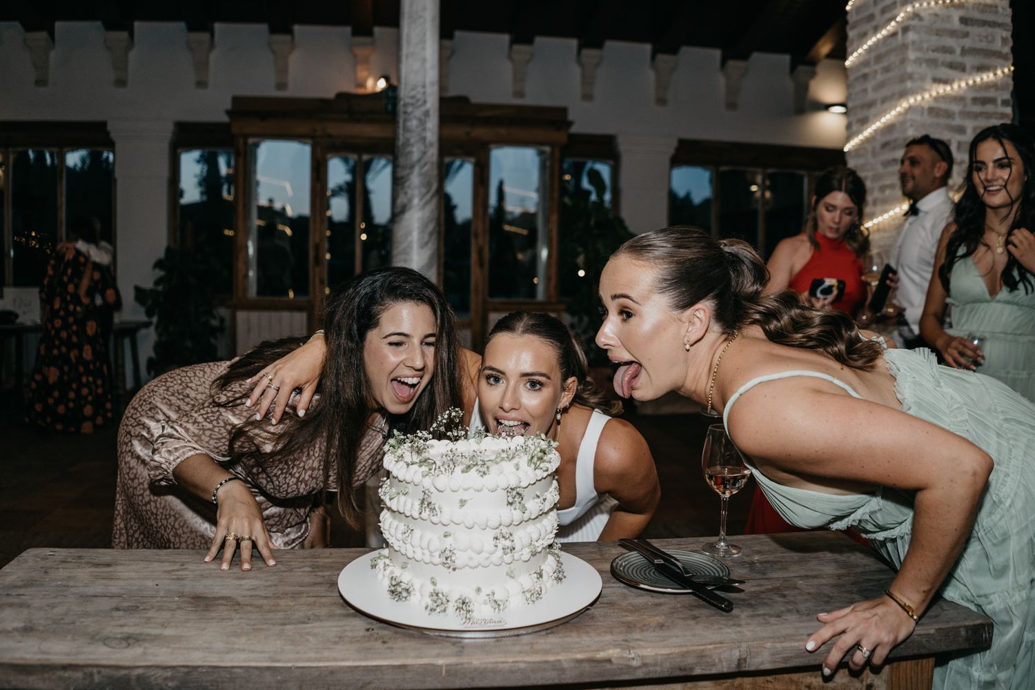 Wedding guests lean in to playfully bite a white tiered wedding cake together at a reception.