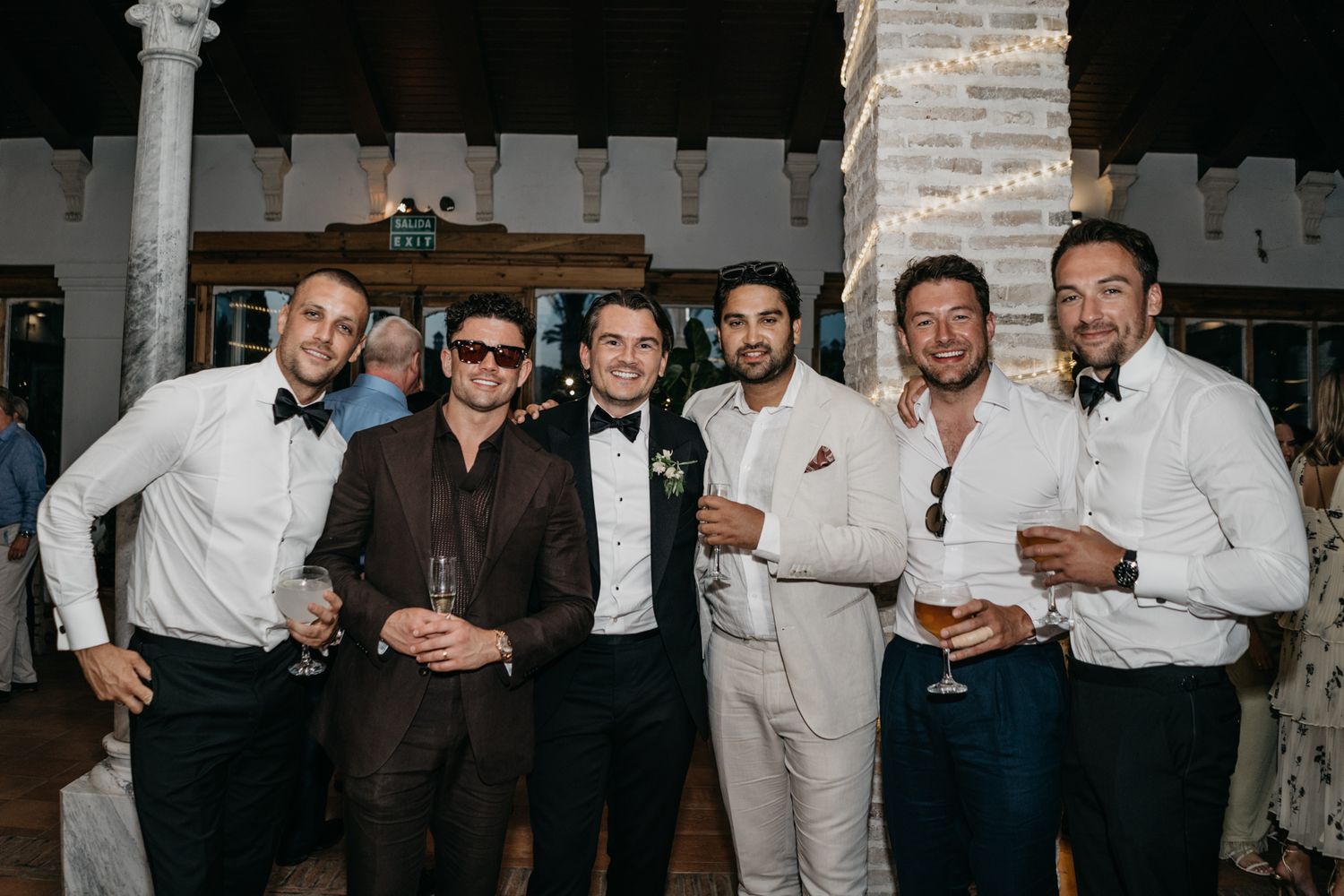 Group of groomsmen in formal attire pose together at an evening wedding reception.