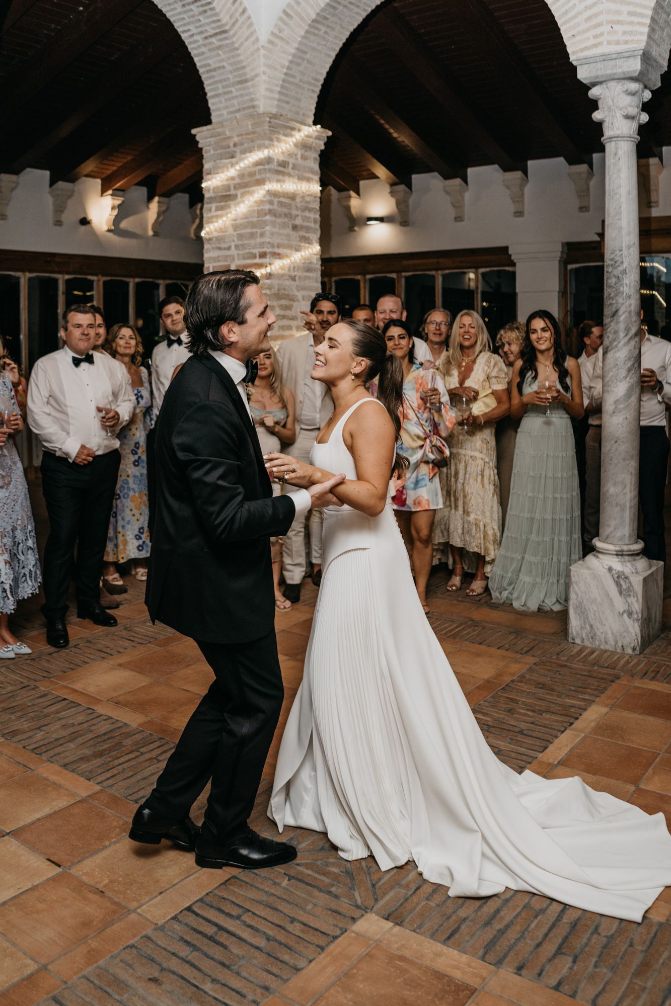 Couple dances together surrounded by wedding guests in an elegant arched venue.